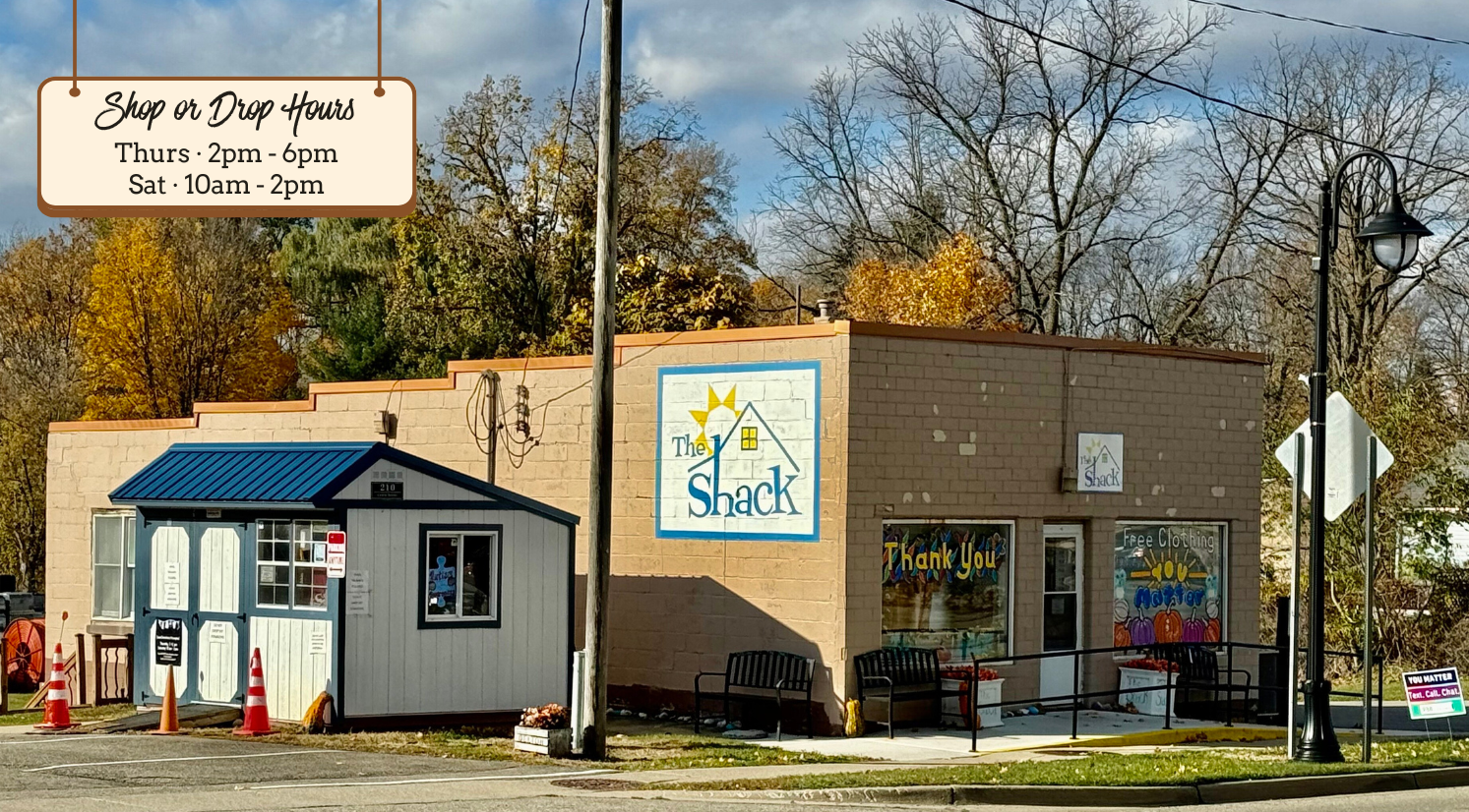 Front view of The Shack, a small free-clothing building with a blue roof and tan brick wall painted with The Shack logo. In the foreground, a hanging sign graphic reads “Shop or Drop Hours: Thurs 2–6pm, Sat 10am–2pm.” Trees with fall colors fill the background.